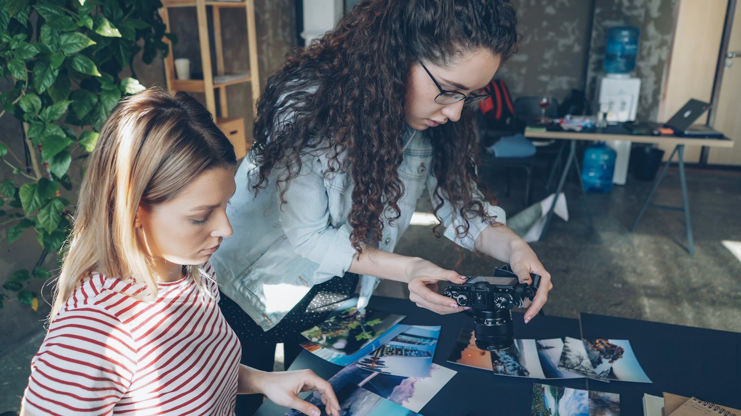 Two women examine photographic prints in a bright creative workspace.